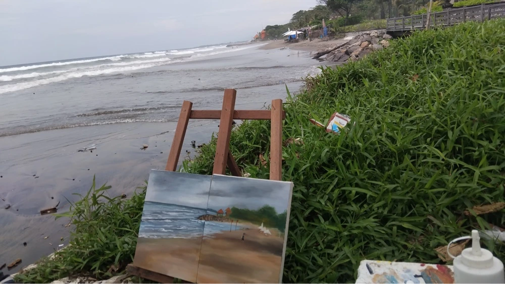 Painting the Spirit of Bali Beneath the Seminyak Sun - Arts of Bali Two landscape paintings displayed on a wooden tripod easel near the shoreline grass at a beach in Kuta