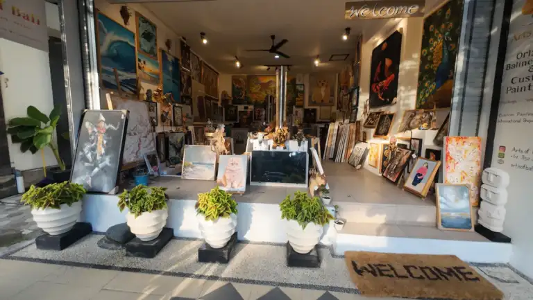 How to Recognize the Real Arts of Bali Gallery in Seminyak - Arts of Bali La galerie d'art « Arts of Bali », située à Seminyak, présente sa façade de jour dans toute sa splendeur, avec ses pots de fleurs blancs, son paillasson de bienvenue et une riche collection de peintures originales visibles à travers la devanture ouverte de la galerie, au n° 42 de la Jl. Raya Seminyak.