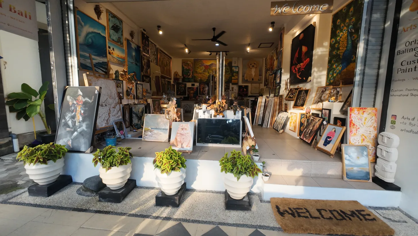 Arts of Bali art gallery in Seminyak showing the full daytime facade with white plant pots, welcome mat, and a rich collection of original paintings visible through the open gallery front on Jl. Raya Seminyak No.42