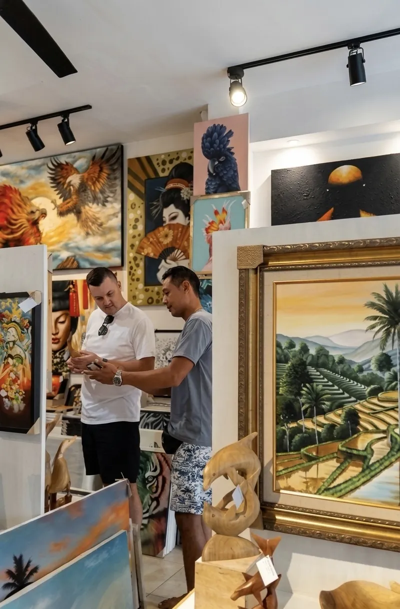 A gallery visitor in a white shirt consulting with an Arts of Bali staff member inside the Seminyak gallery, with a large gold-framed Balinese landscape painting by Gandara and diverse artworks visible on the walls behind them