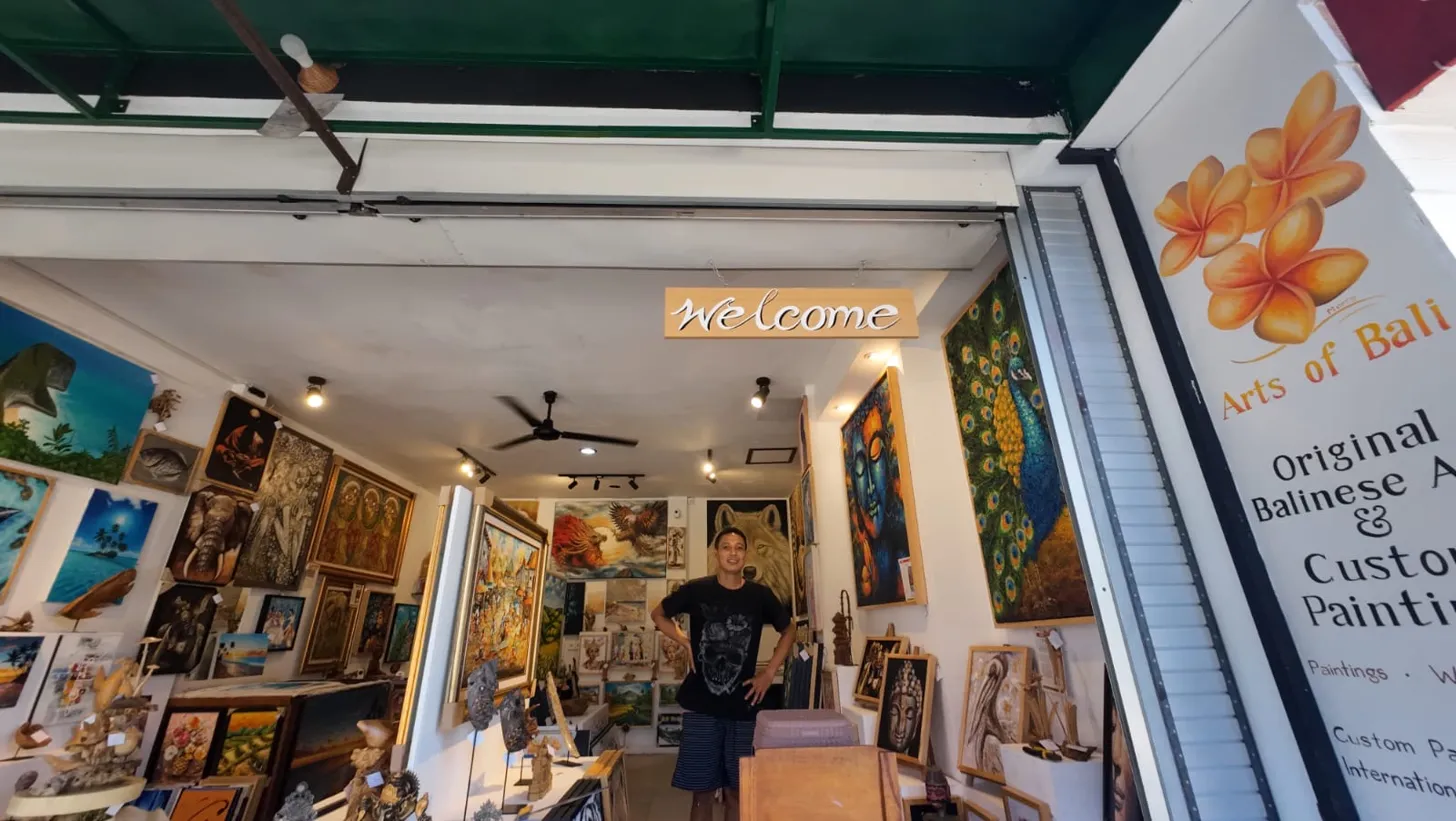 Putu, owner of Arts of Bali gallery, standing at the gallery entrance in Seminyak under a welcome sign with original paintings and the Arts of Bali banner visible behind him