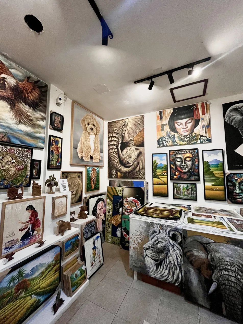 Interior of Arts of Bali art gallery Seminyak showing a wall display of original paintings including a hyperrealist dog portrait by Alzen, elephant, Balinese woman, Buddha, and landscape works alongside wooden sculptures