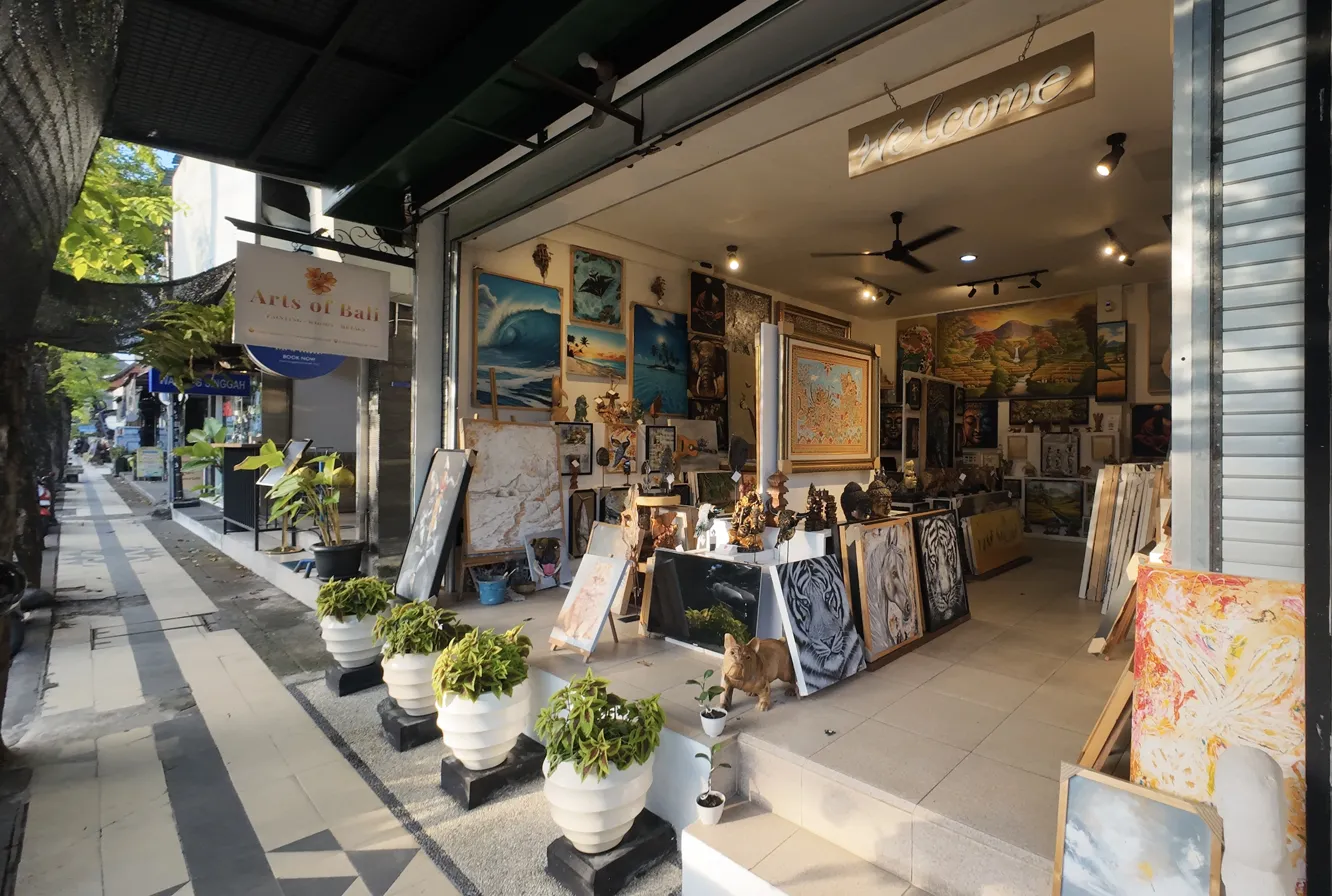 Exterior view of Arts of Bali art gallery Seminyak showing the gallery entrance with Arts of Bali signboard, welcome sign, diverse original paintings displayed at the front, and the Jl. Raya Seminyak sidewalk