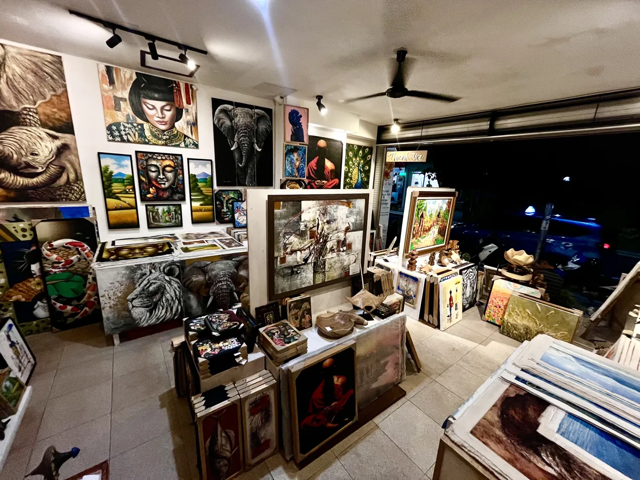 Night-time interior of Arts of Bali gallery in Seminyak showing the full collection illuminated under gallery lighting, with paintings covering every wall from floor to ceiling and Jl. Raya Seminyak visible through the open gallery front