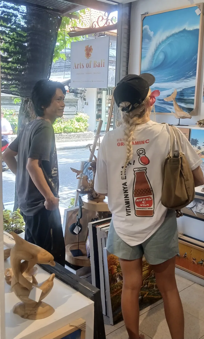 A female visitor browsing at Arts of Bali gallery in Seminyak with a staff member, the Arts of Bali sign clearly visible in the background alongside original ocean and coastal paintings on the gallery walls