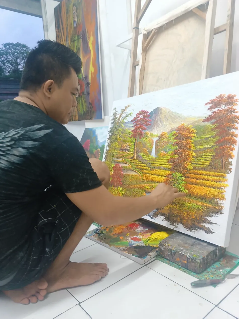 Balinese landscape painter sitting on the studio floor adding finishing detail to a large bali rice field painting on canvas with paint palette beside him at Arts of Bali studio Seminyak