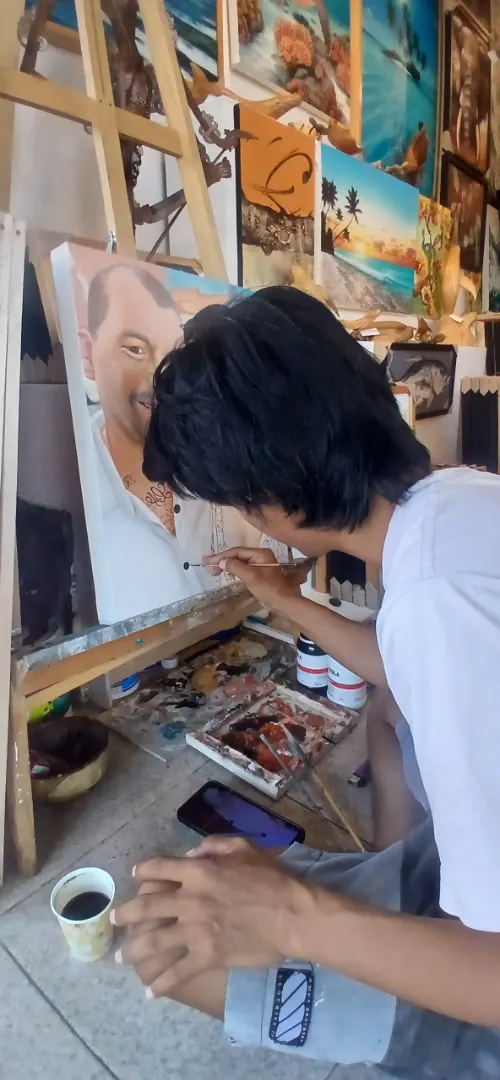 Balinese portrait artist Alzen leaning close to the canvas to paint fine tattoo detail with a thin brush during the finishing stage of a custom portrait commission at Arts of Bali in Seminyak