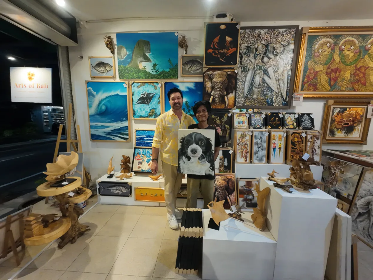 A collector holding a custom hyperrealist dog portrait painting inside Arts of Bali gallery in Seminyak, with diverse original artworks visible on the walls behind them