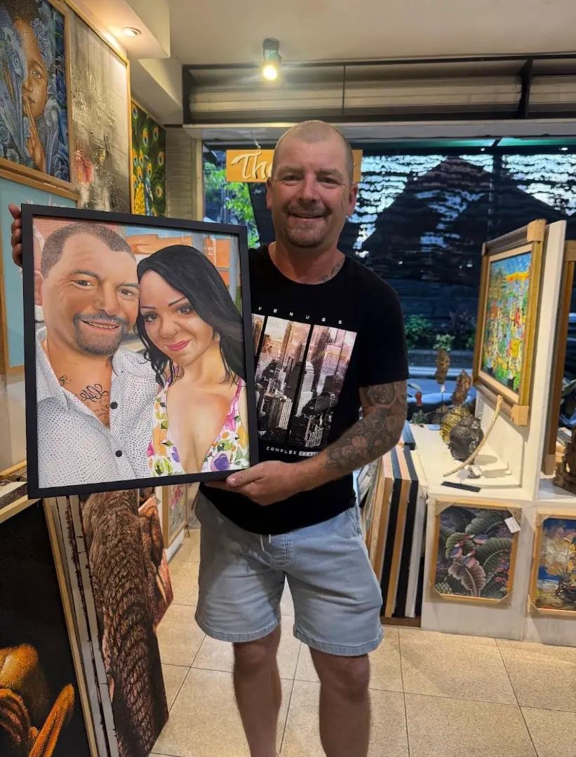 A happy Australian client holding his finished commissioned portrait painting inside Arts of Bali gallery in Seminyak, smiling as he sees the completed oil painting of himself and his partner for the first time