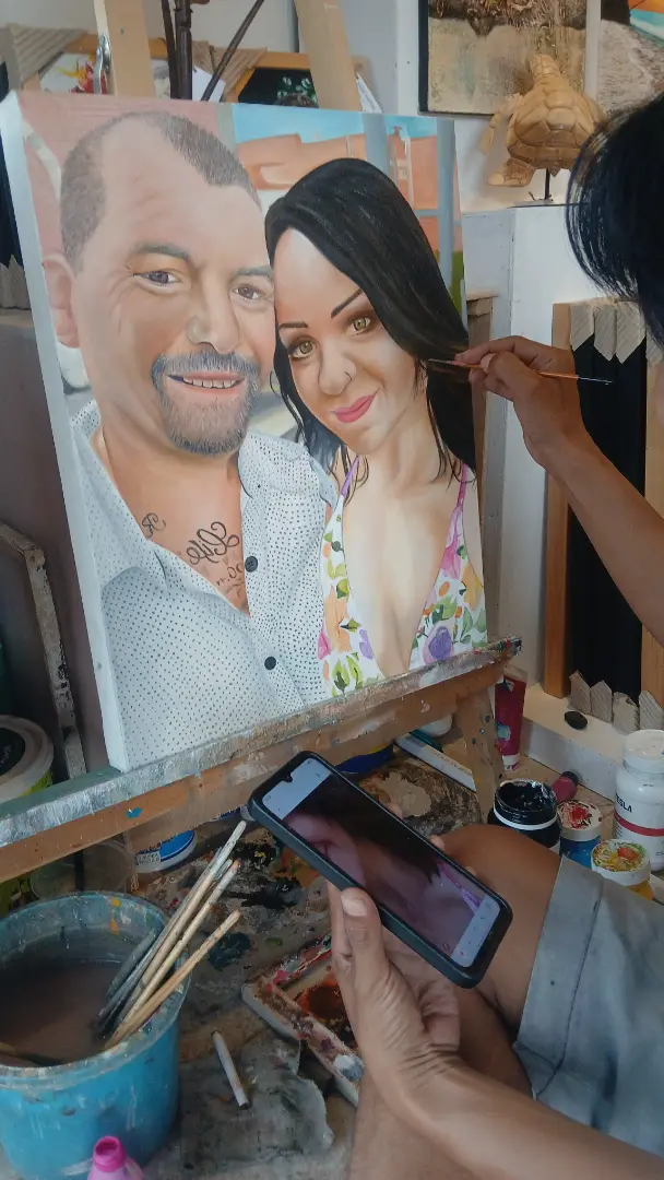 Arts of Bali portrait artist Alzen adding final details to a near-complete custom couple portrait while holding the reference photo on a phone for comparison — both subjects fully painted in oil on canvas
