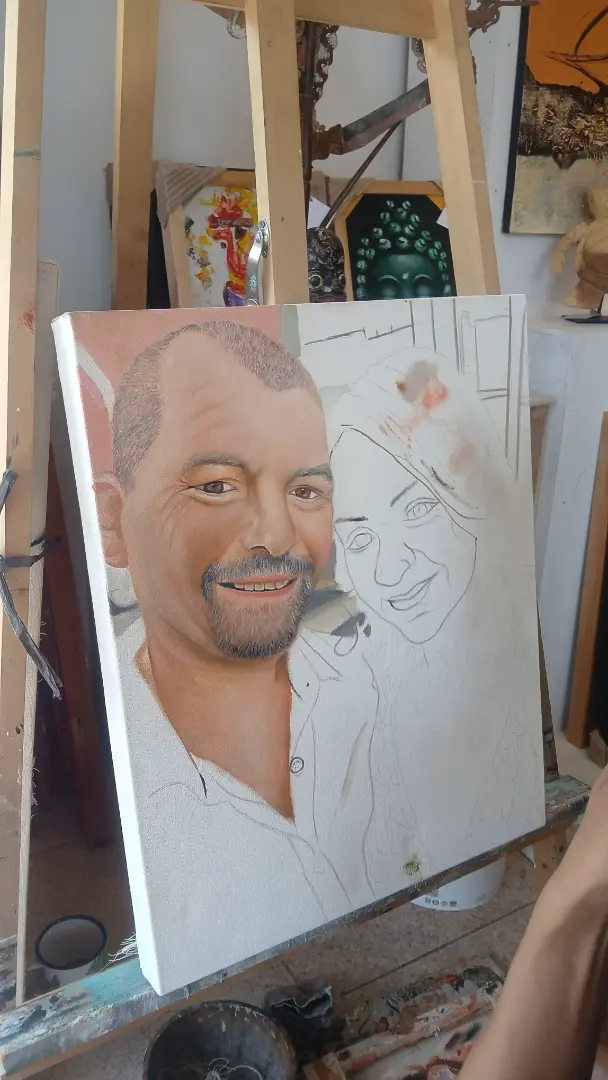 A custom portrait painting in Bali at the halfway point — the man is fully rendered in oil paint with skin, beard and tattoo detail complete, while the woman beside him remains as a pencil sketch on the same canvas at Arts of Bali
