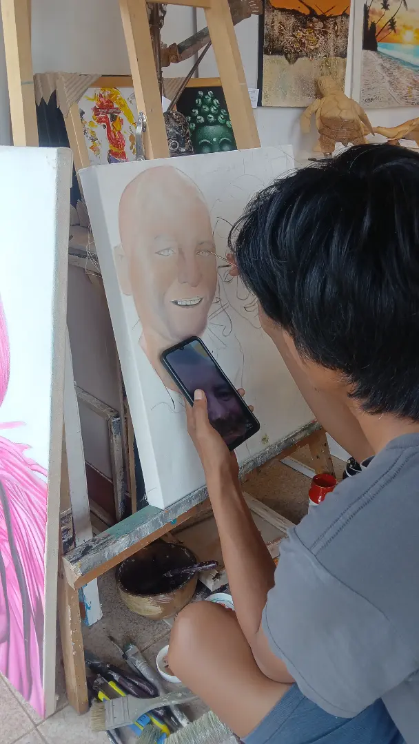 A Bali portrait artist holding a phone next to the canvas to compare the reference photo while painting the early skin tone layers of a custom oil portrait commission at Arts of Bali