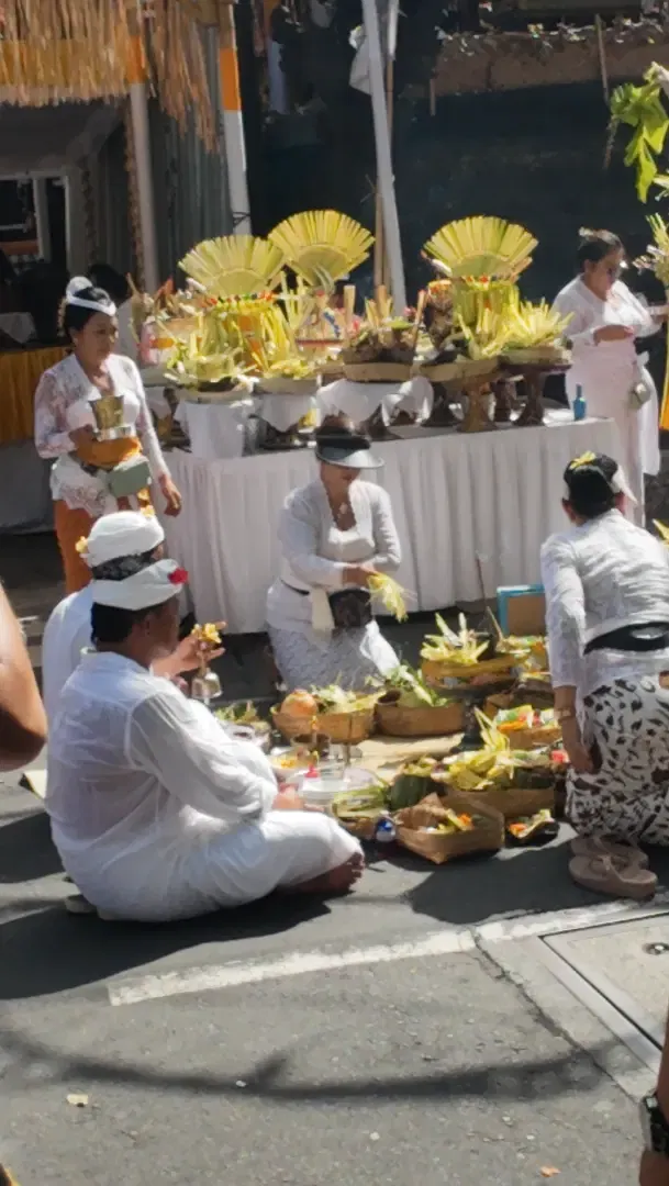 Balinese women in white ceremonial dress seated on the ground carefully arranging and sequencing banten offerings in woven baskets on a white-clothed table during Odalan ceremony preparation in Seminyak Bali
