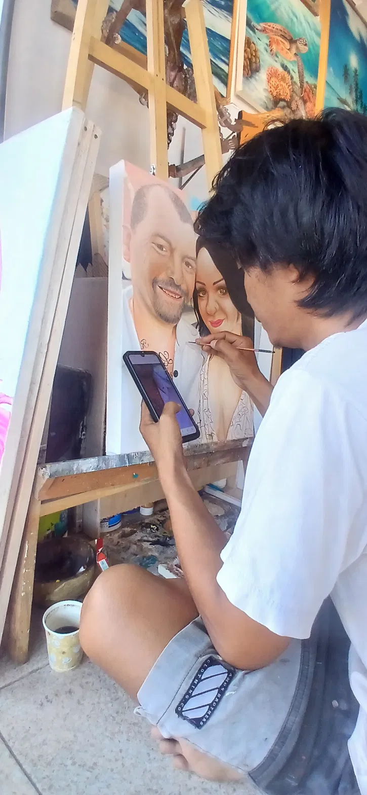 Portrait Specialist Referencing Client Photo While Painting Custom Couple Portrait Arts of Bali portrait specialist seated at the easel, referencing the client's original photo on his phone while painting a large hyper-realistic custom couple portrait