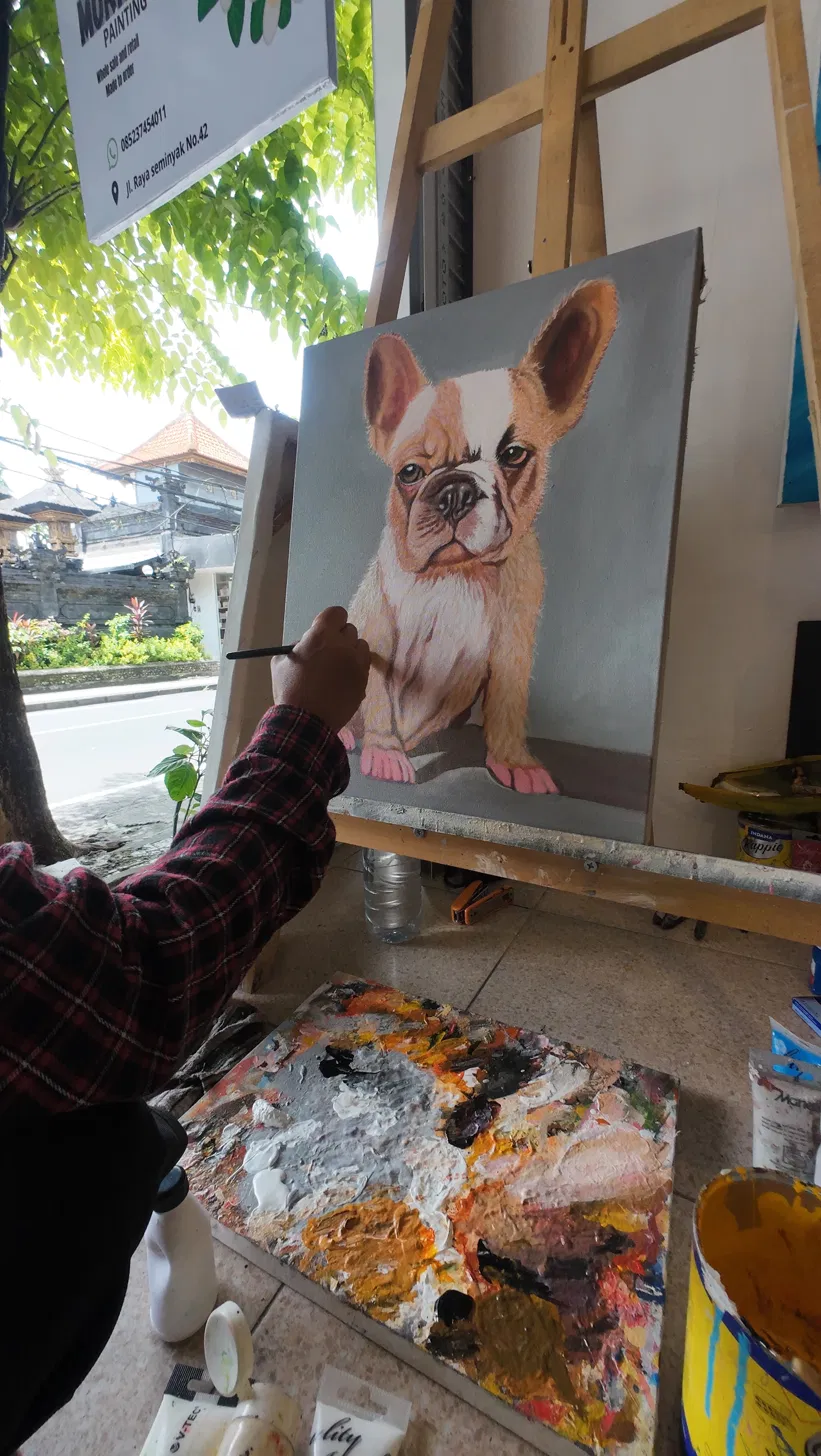 Hyper-Realistic French Bulldog Pet Portrait in Progress at Arts of Bali Seminyak Studio Arts of Bali artist carefully adding fine detail to a hyper-realistic French Bulldog pet portrait on an easel, with a heavily-loaded colour palette visible in the foreground