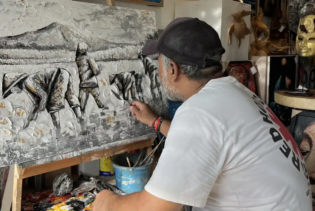 Balinese artist Upeksa working in studio at Arts of Bali Seminyak — applying palette knife to monochrome black and white impasto painting showing farmers, palette knife technique visible