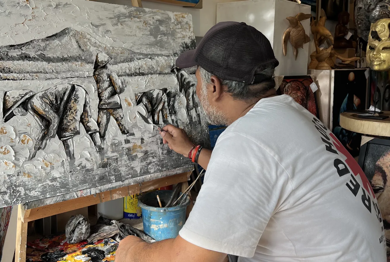 Balinese artist Upeksa working in studio at Arts of Bali Seminyak — applying palette knife to monochrome black and white impasto painting showing farmers, palette knife technique visible