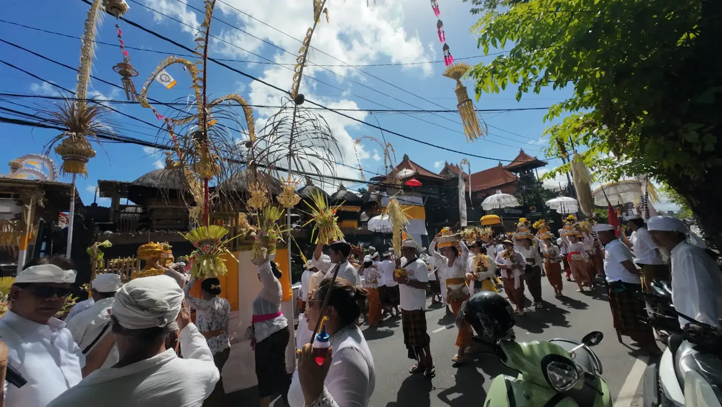 Large Balinese Odalan ceremony procession filling Jl. Raya Seminyak with hundreds of worshippers in white carrying banten offerings and decorated penjor bamboo poles under a clear blue sky, photographed 26 May 2026