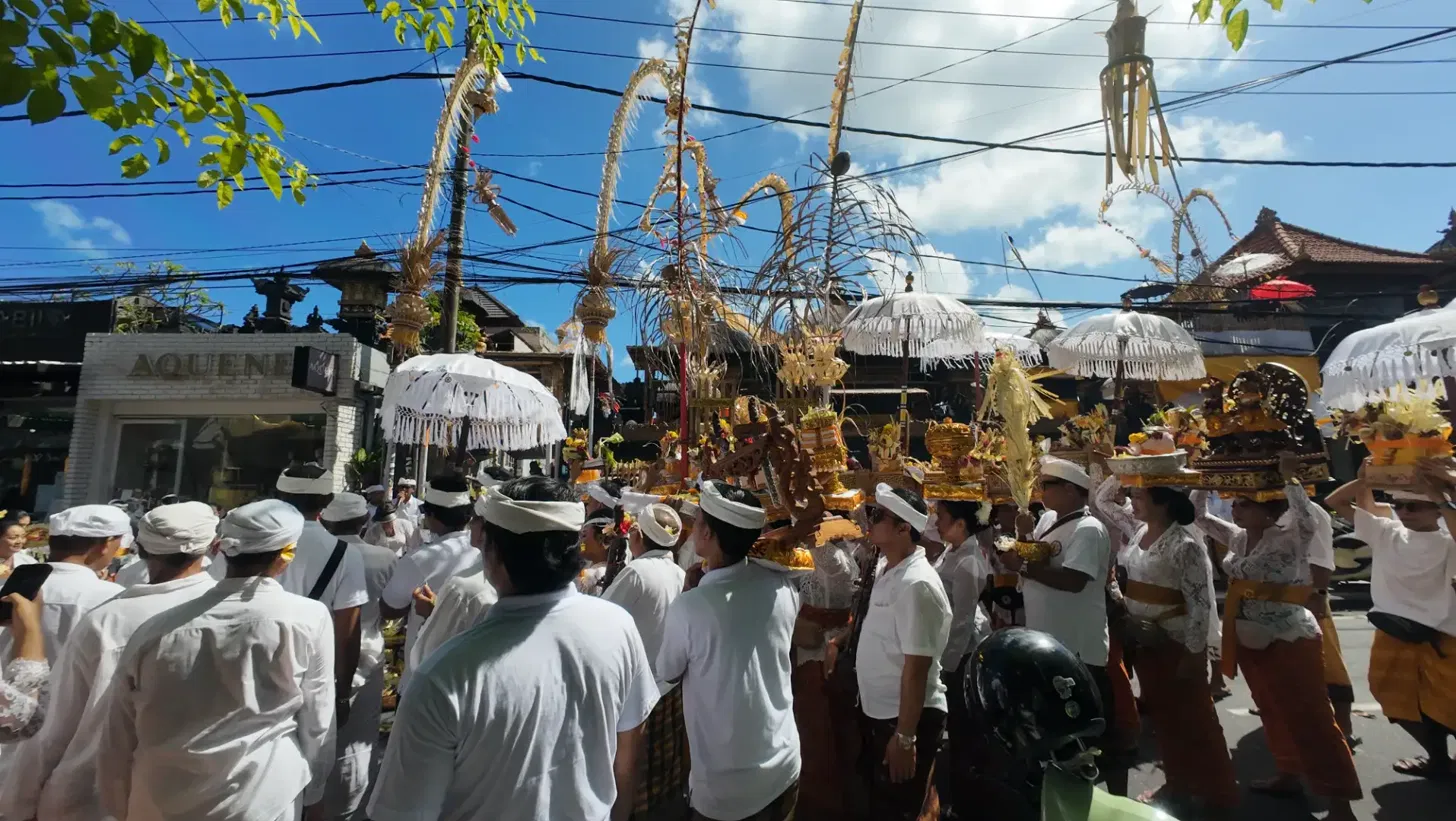 Dense crowd of Balinese worshippers in white ceremonial dress carrying banten offerings on their heads under rows of white tedung ceremonial umbrellas during an Odalan procession on Jl. Raya Seminyak Bali