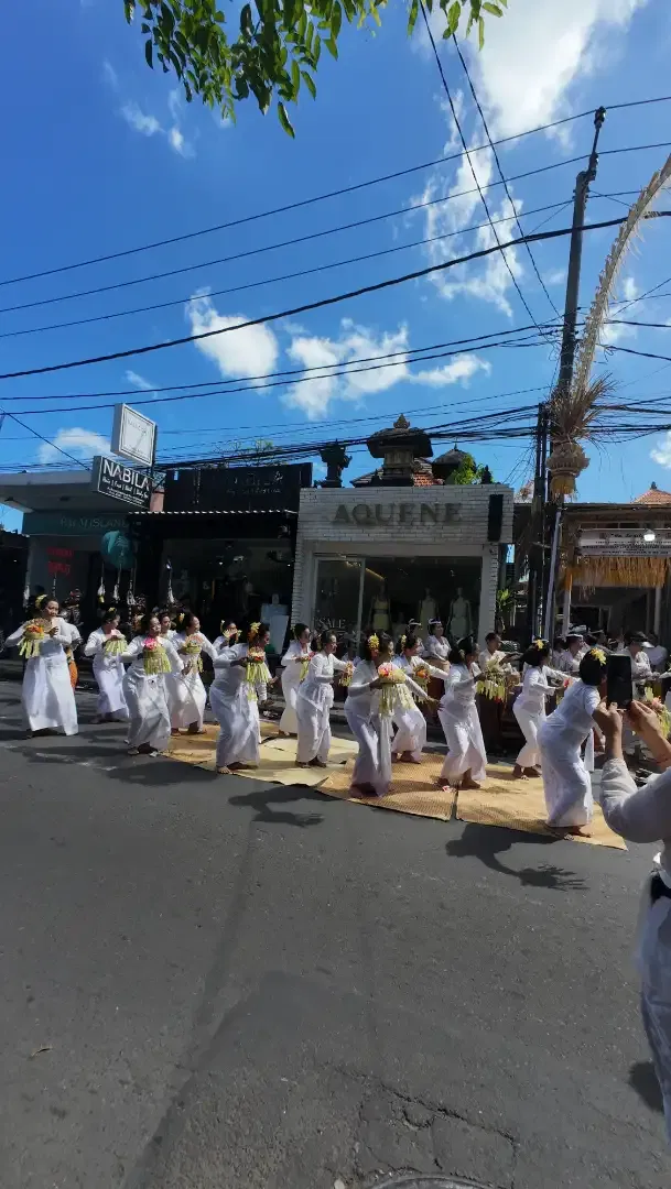 A long line of young Balinese women in white kebaya performing the Rejang sacred offering dance in perfect formation on the street in front of Seminyak shopfronts during an Odalan ceremony, 26 May 2026