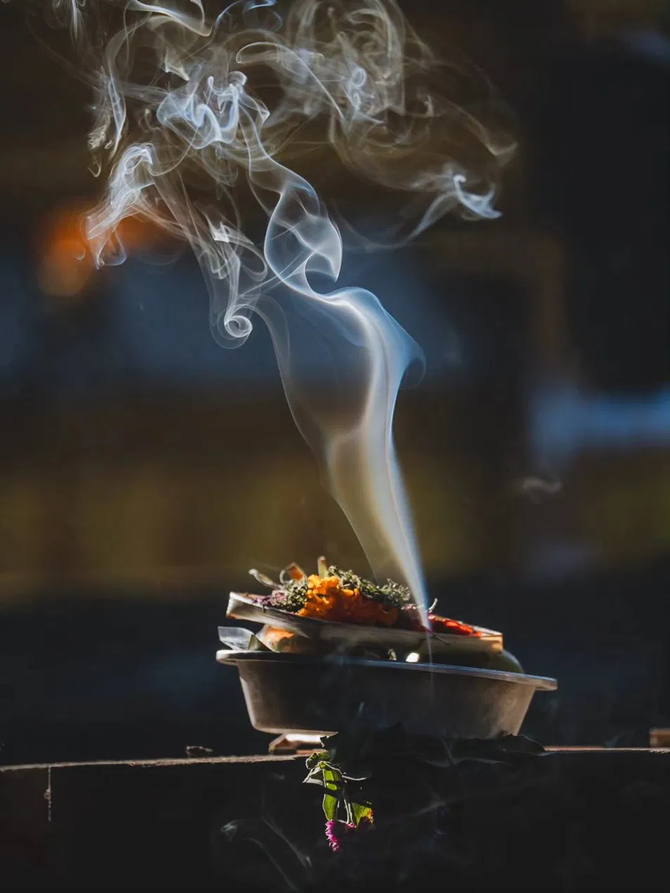 Smoke rising in delicate curling patterns from a canang sari offering with marigold flowers and incense, backlit against a dark background, small Balinese temple offering tray