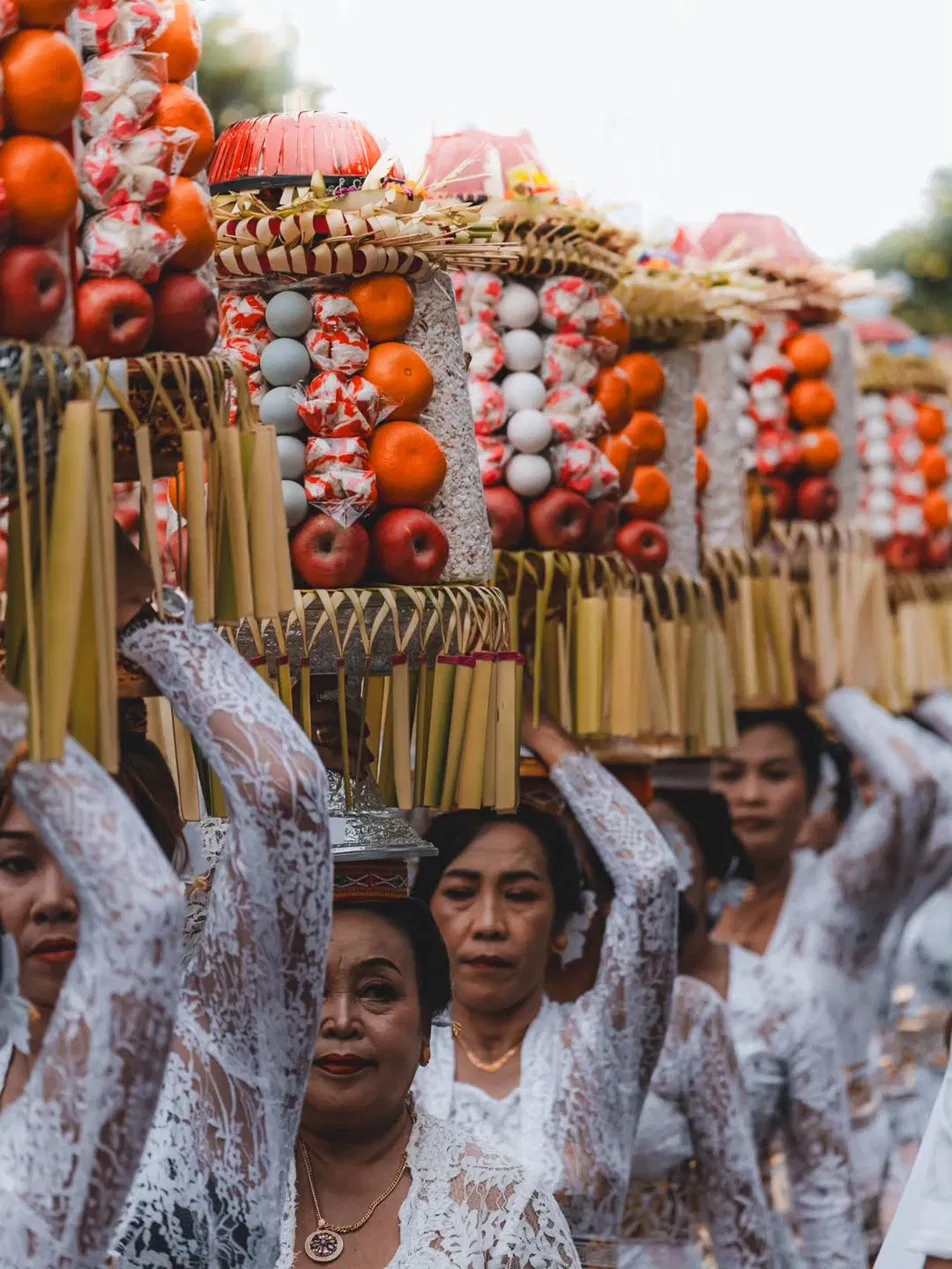 Close-up of Balinese women in white lace kebaya carrying tall gebogan fruit offering towers on their heads in procession, oranges apples and rice cakes visible, decorative woven palm leaf trim