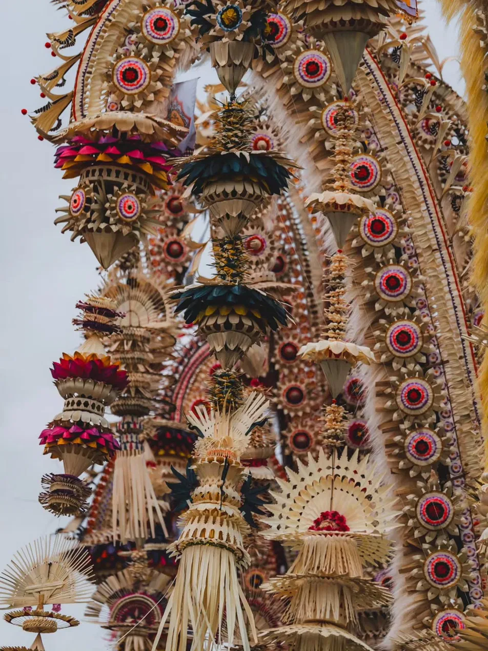 Close-up detail of penjor sampian decorations during Galungan in Bali, intricately woven palm leaf forms in red, gold, purple, and cream, layered cone and fan shapes hanging against a pale sky