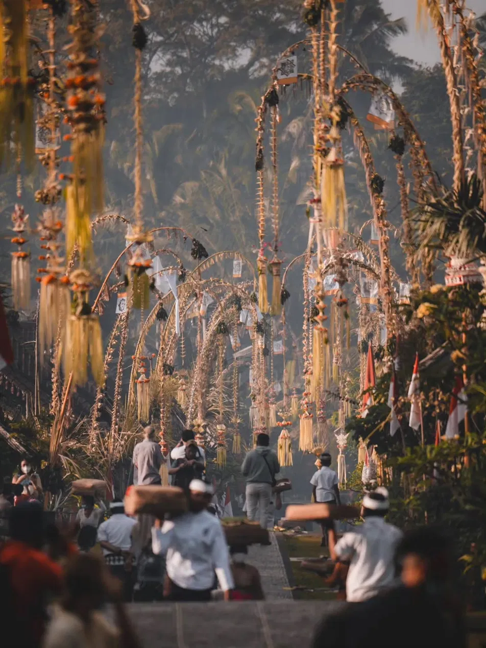 Misty morning street in Bali during Galungan lined with tall curving penjor bamboo poles, people walking in procession carrying offerings on their shoulders and heads toward a temple, golden light filtering through trees