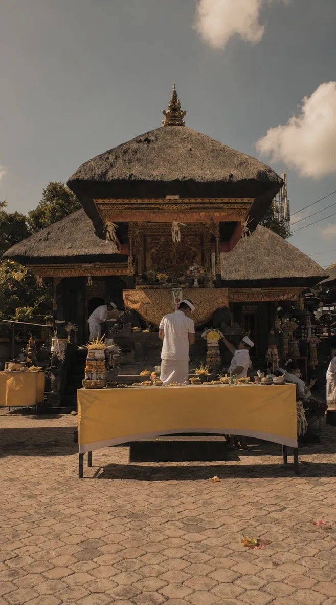 Tall thatched-roof Balinese temple shrine with gold decorative carving, people in white ceremonial dress preparing offerings on a long table covered in yellow cloth in the foreground, clear blue sky