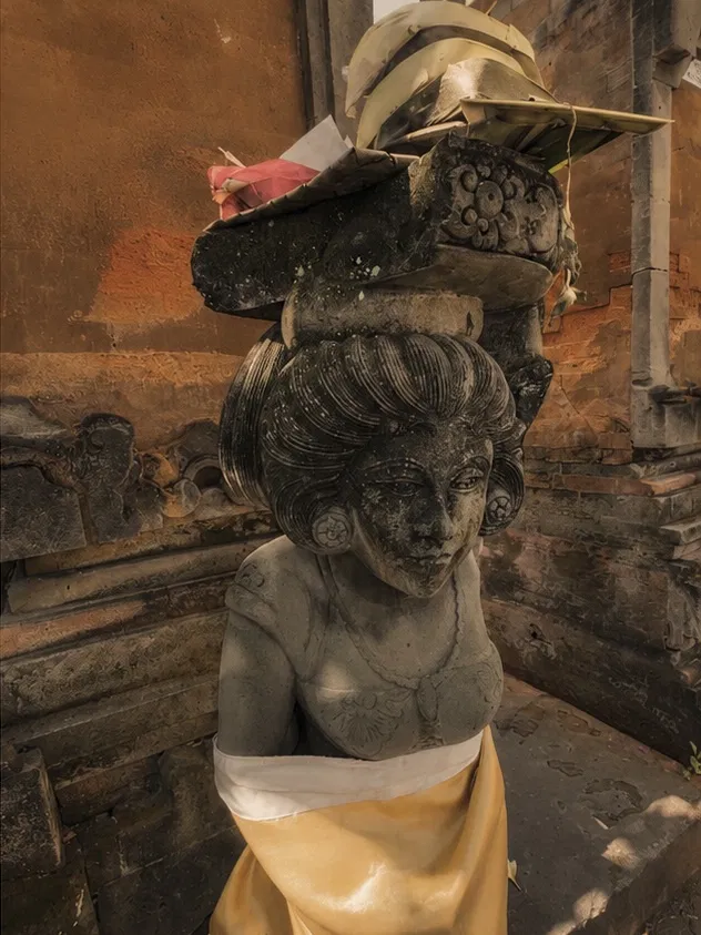 Stone statue of a Balinese woman in the suun pose, carrying a tall offering on her head, weathered dark patina, white and gold cloth wrapped at the base, temple wall background