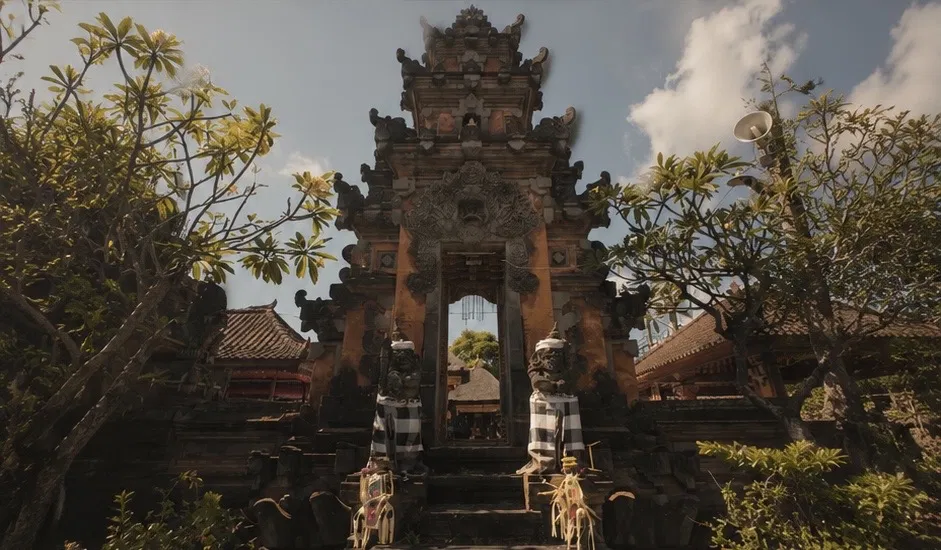 Candi bentar split temple gate in Bali flanked by two stone guardian statues wrapped in black and white poleng cloth, frangipani trees on either side, blue sky with clouds, temple roofs visible through the gate
