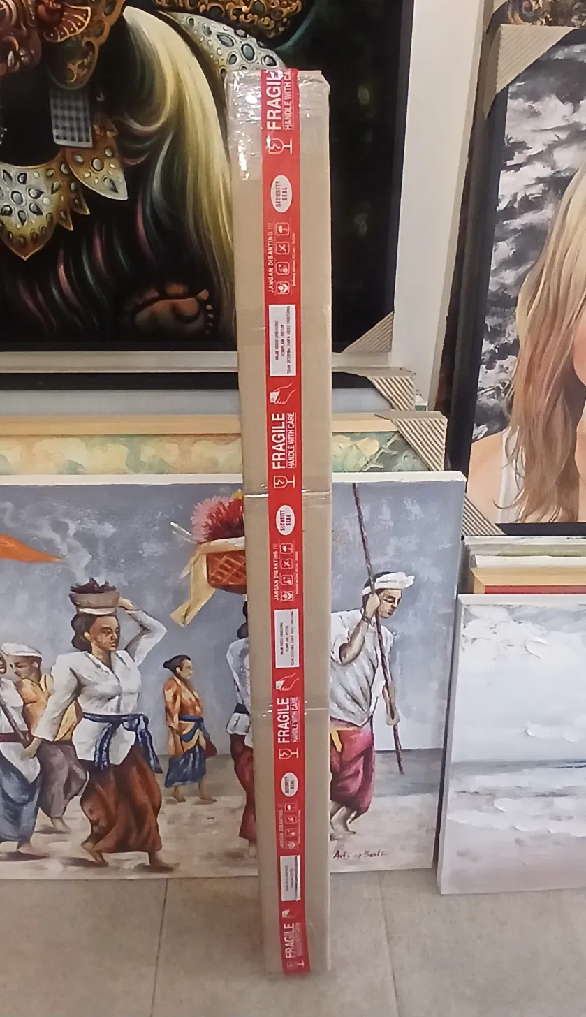 Long cardboard shipping tube wrapped in red FRAGILE HANDLE WITH CARE tape and security seals, leaning against framed Balinese paintings inside Arts of Bali gallery before being sent for international shipping