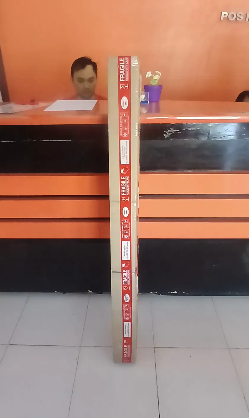 Long cardboard tube wrapped in red fragile tape standing upright at a Pos Indonesia post office counter, ready for international shipping, orange counter and tiled floor visible