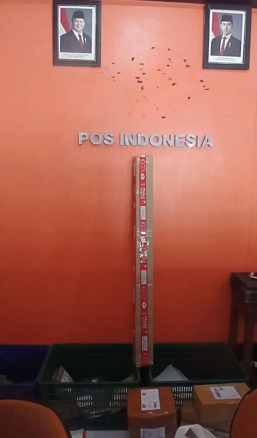 Rolled canvas package wrapped in red fragile tape standing against an orange wall with POS INDONESIA signage, prepared for international art shipment from Bali