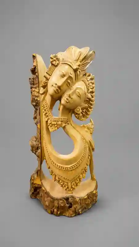 Balinese wood carving of pengantin couple in tall elongated vertical form, two faces leaning toward each other with elaborate interlocking headdresses, detailed jewelry at neck and chest, grey background, Arts of Bali Seminyak