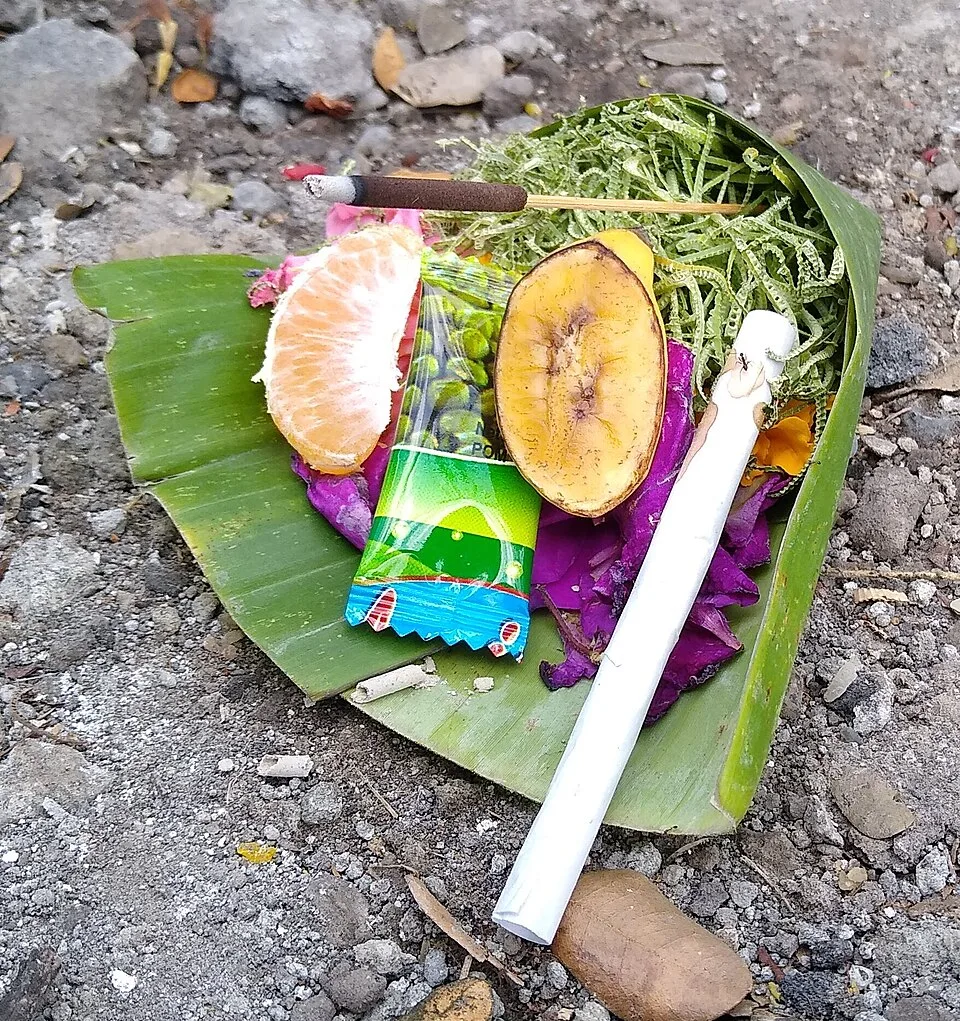 A canang sari in Canggu on banana leaf with a mandarin segment, a snack packet, petals and an incense stick