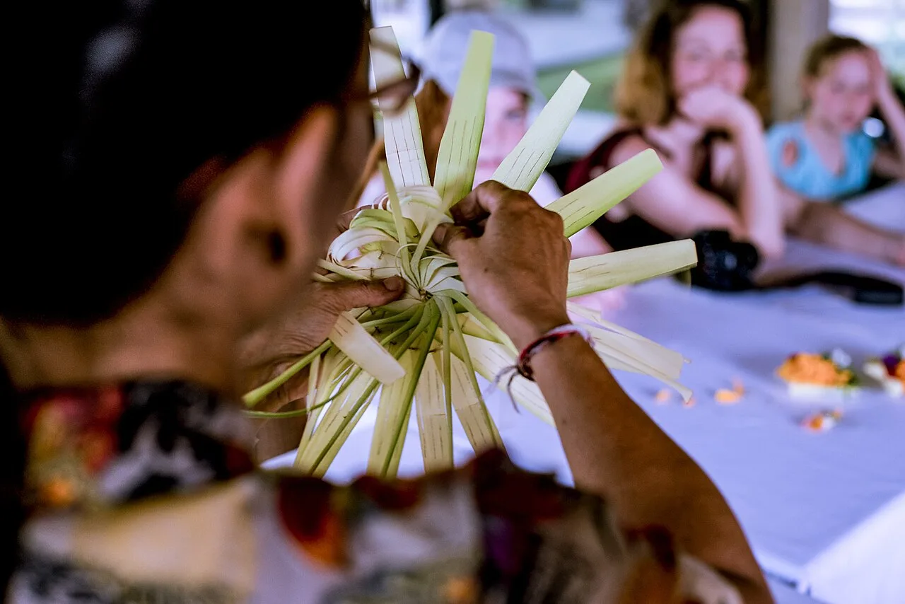 Hands folding strips of young coconut leaf while a Balinese woman teaches tourists to make a canang