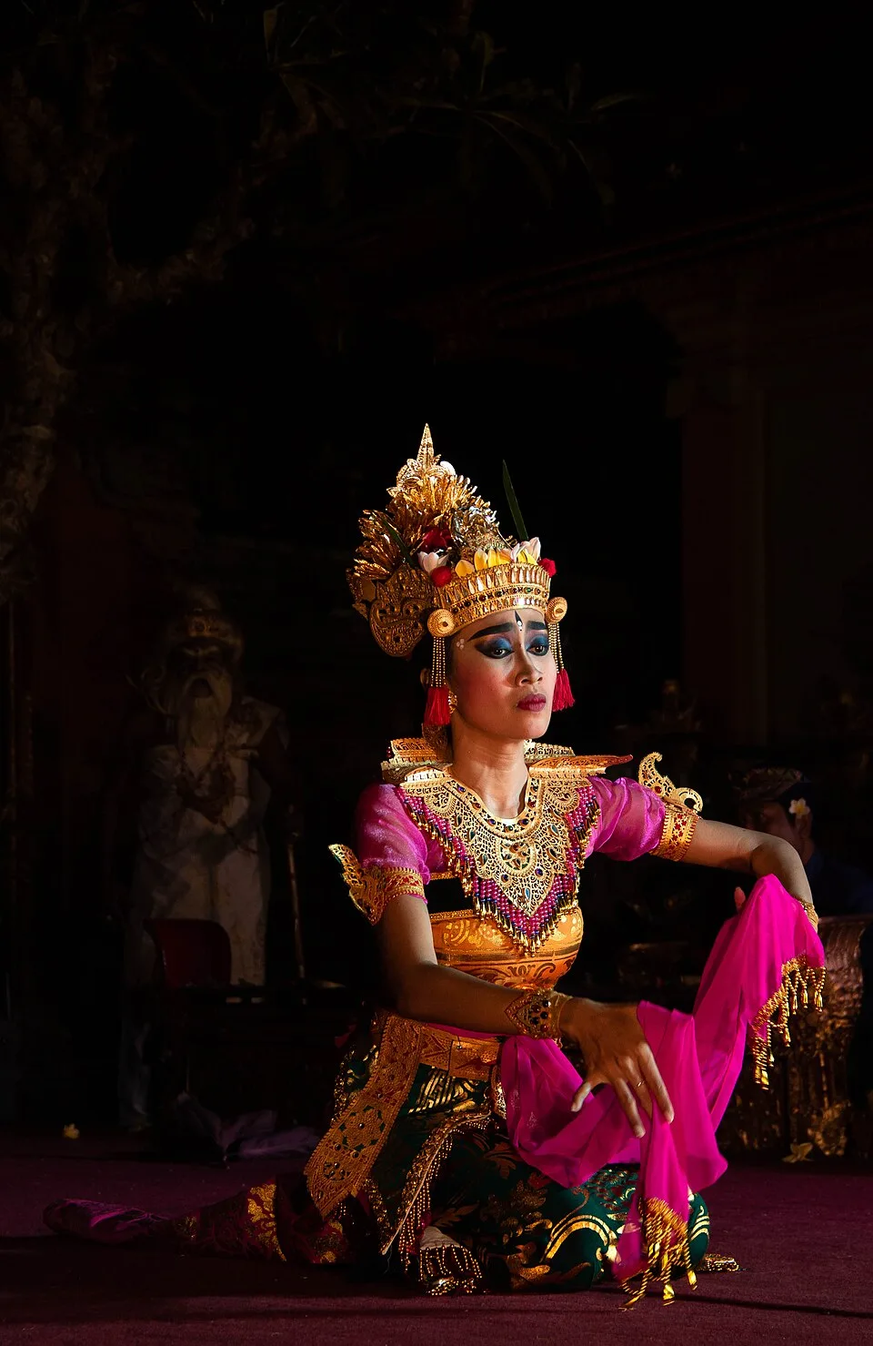 Legong dance performer in a gold crown and pink and green costume kneeling on stage at Ubud Palace