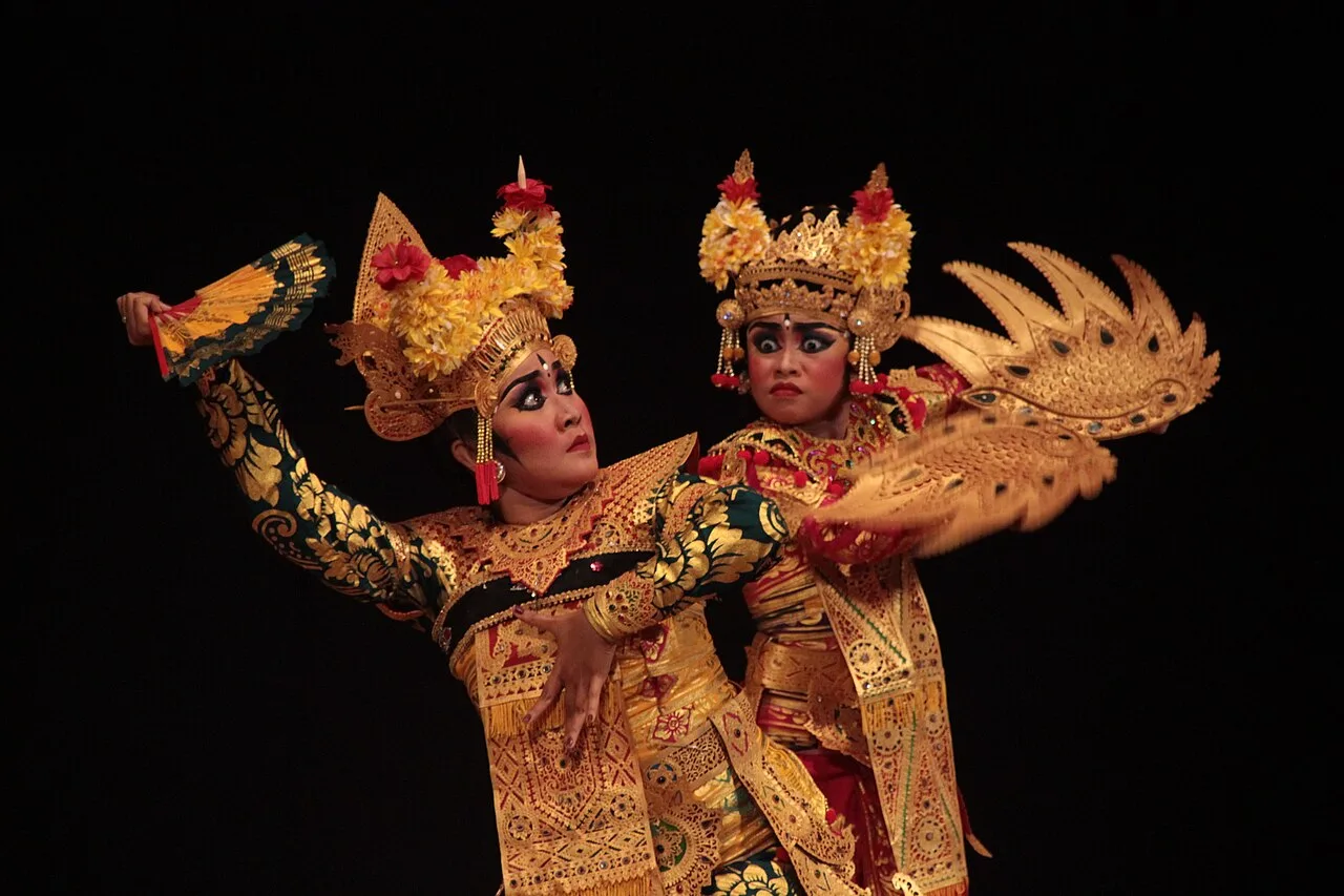 Two legong dance performers in gold costumes and flower headdresses holding fans in an agem pose