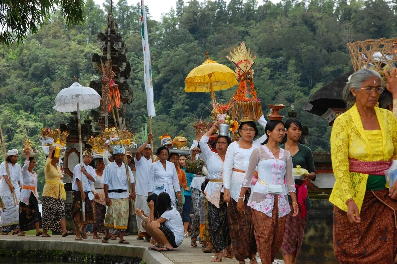 Worshippers carrying offerings on their heads in a procession toward a Balinese temple gate, under yellow and white umbrellas