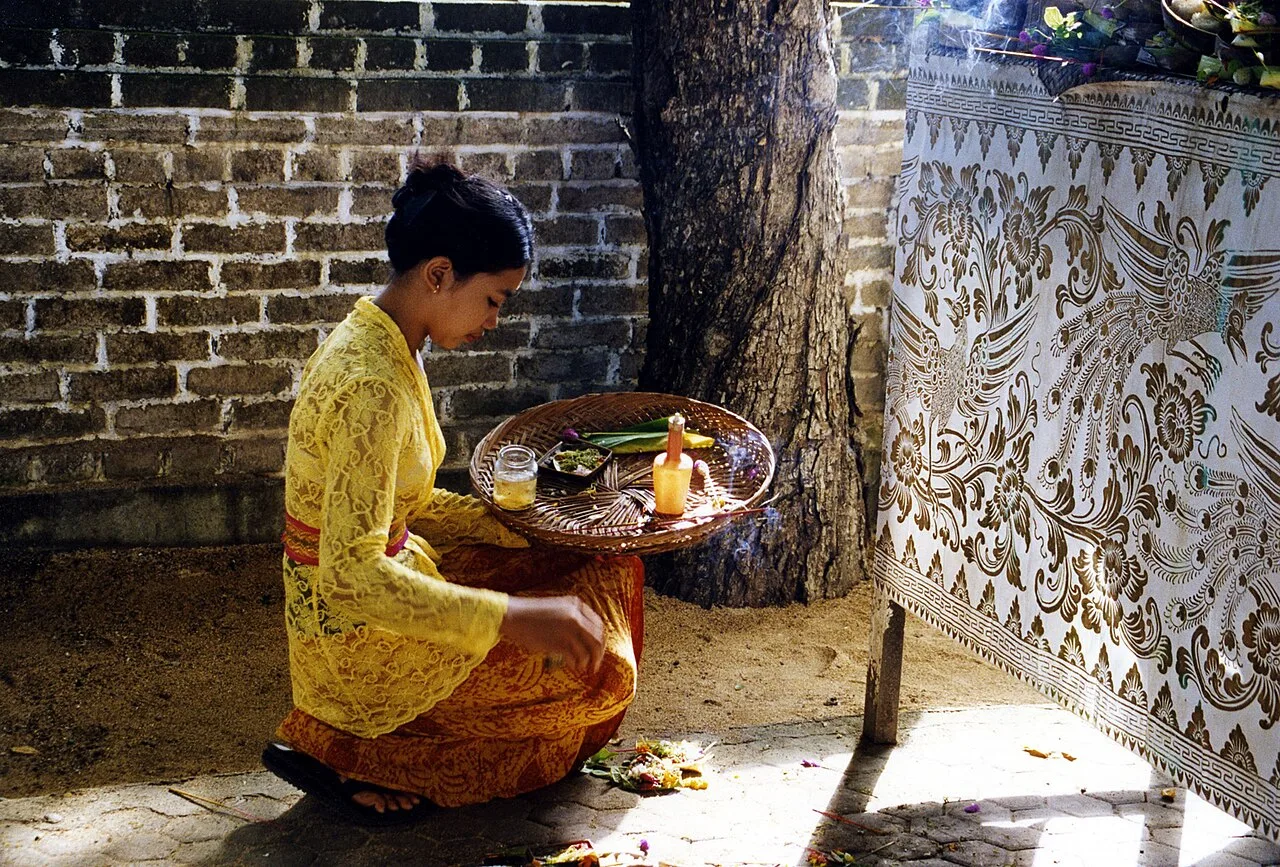 Balinese woman in a yellow blouse holding an offering tray beside a brick wall and a tree